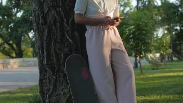 Krullende vrouw met skateboard is met behulp van mobiele telefoon in het openbaar park. — Stockvideo