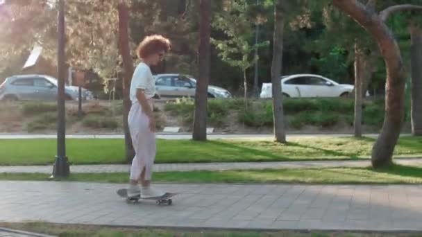 Mooi jong meisje met krullend haar paardrijden op skateboard tijdens zonsondergang in het park — Stockvideo