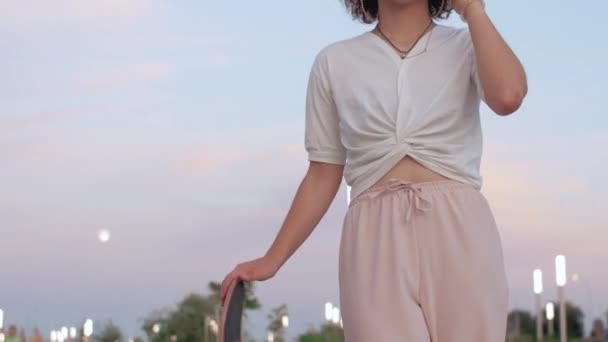 Curly woman with skateboard is talking on the phone In The Public Park. — Stock Video