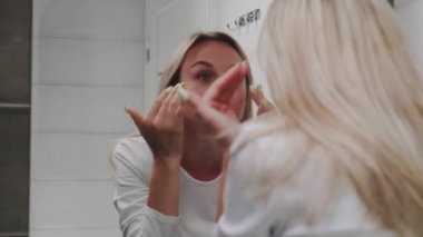 Banyoda aynanın önünde beyaz bir kadın var. Yüz yıkama, cilt bakımı, sağlık.