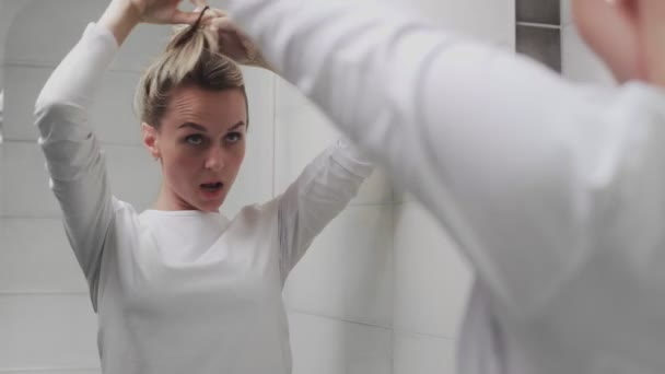 Uma jovem mulher enfia o cabelo em uma faixa elástica e olha para o espelho — Vídeo de Stock