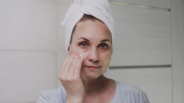 Взрослая женщина с полотенцем на голове наносит крем и смотрит в камеру — стоковое видео