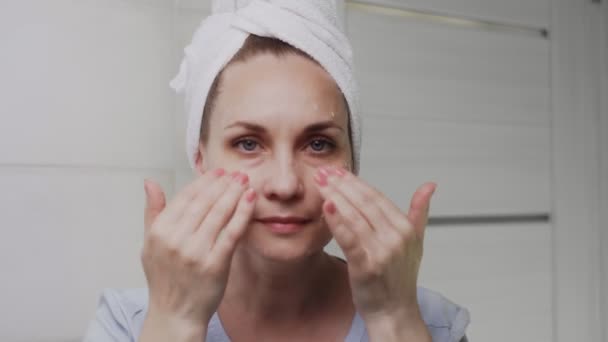 Adult woman with a towel on her head applying cream and looking at the camera — Stock Video