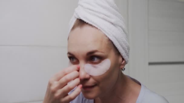 Крупным планом взрослая женщина с полотенцем на голове удаляет коллагеновые прокладки под глазами и смотрит в зеркало — стоковое видео