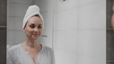 Banyoda aynanın önünde kafasında havlu olan beyaz bir kadın var. Yüz yıkama, cilt bakımı, sağlık.
