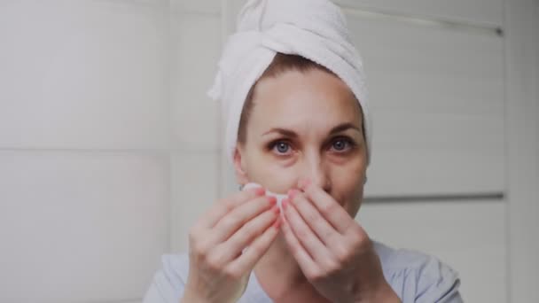 Крупным планом взрослая женщина с полотенцем на голове кладет коллагеновые прокладки под глаза и смотрит в камеру — стоковое видео