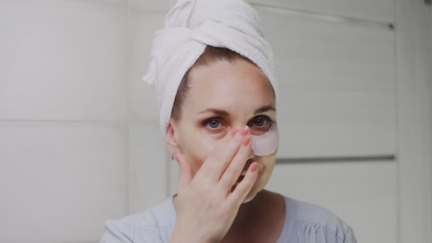 Gros plan de la femme adulte avec une serviette sur la tête enlève les tampons de collagène sous ses yeux et regarde la caméra — Video