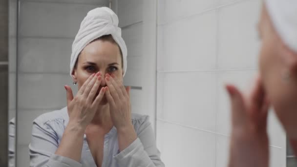 Frusztrált felnőtt nő törülközővel a fején ránéz az arcára a fürdőszobában tükörben. — Stock videók
