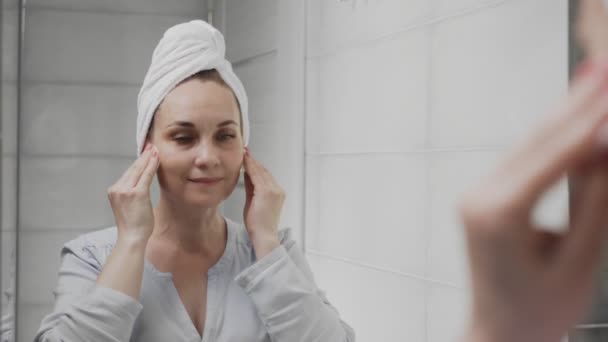 Mujer adulta con una toalla en la cabeza aplicando loción y mirando en el espejo disfrutar de la belleza natural y saludable — Vídeos de Stock