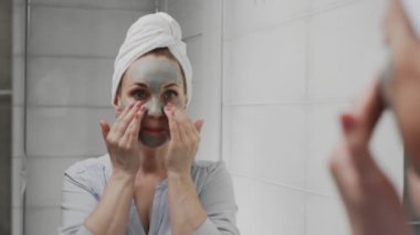 Kafasında havlu olan yetişkin bir kadın kameraya bakarak yüzüne kilden maske takıyor.