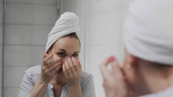 Μια ενήλικη γυναίκα με πετσέτα στο κεφάλι βάζει κολλαγόνο κάτω από τα μάτια της και κοιτά στον καθρέφτη. — Αρχείο Βίντεο