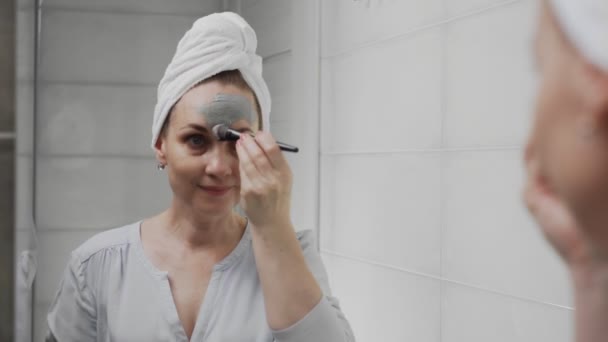 Mujer adulta con una toalla en la cabeza aplicando máscara de arcilla en su cara mirando a la cámara — Vídeos de Stock