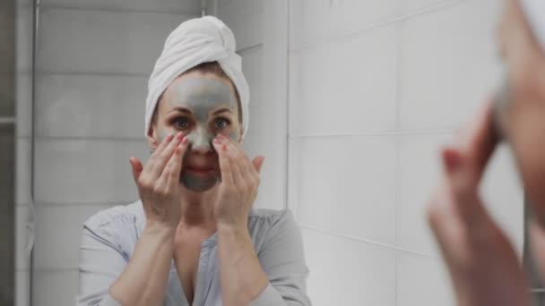 Felnőtt nő törülközővel a fején, agyagmaszkkal az arcán, ahogy a kamerába néz. — Stock videók
