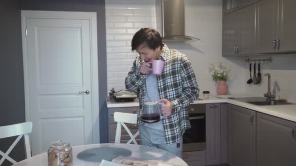 Νεαρός Ασιάτης χασμουριέται και πίνει καφέ στην κουζίνα στο σπίτι — Αρχείο Βίντεο