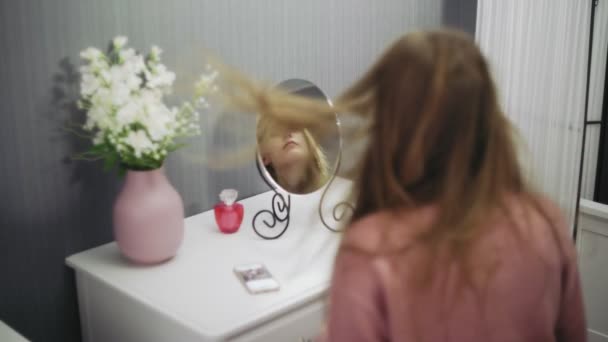 Hermosa mujer joven está utilizando un secador de pelo mientras se mira en el espejo en la habitación — Vídeos de Stock