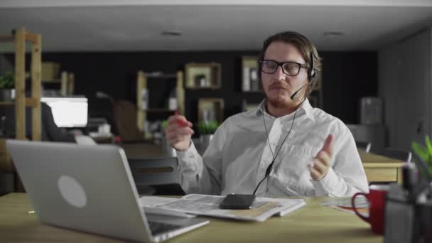 Joven gerente de auriculares está trabajando en el ordenador portátil en la oficina — Vídeos de Stock