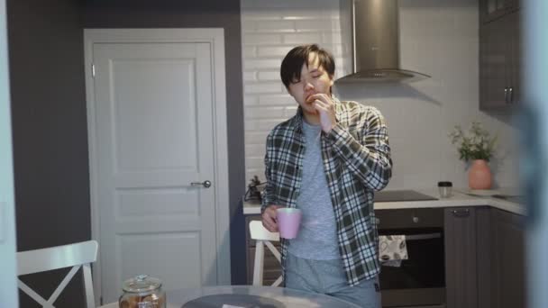 Young sick asian man eating cookie and drinking coffee in kitchen at home — Stock Video
