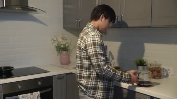 Sleepy asian man pouring coffee in cup and taking a cookie at home in the kitchen. — Stock Video