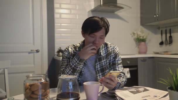 Счастливый азиатский человек читает что-то смешное на смартфоне и улыбается на кухне дома — стоковое видео