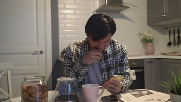 Boldog ázsiai férfi olvas valami vicces okostelefon és nevet a konyhában otthon — Stock videók