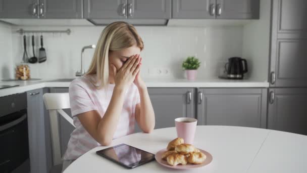 Sad woman with tablet, cup of coffee and croissants on her kitchen — Stock Video