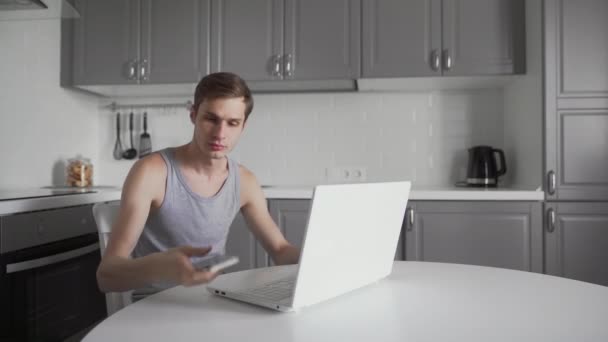Mladý podnikatel mluví na mobilním telefonu a pracuje na notebooku v kuchyni. — Stock video