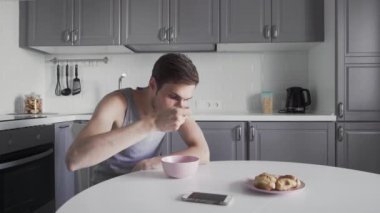 Genç adam çorba içiyor ve mutfakta telefon kullanıyor.