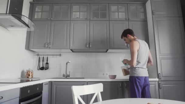 Unge man lagar frukost i köket. han häller majsflingor i en skål — Stockvideo