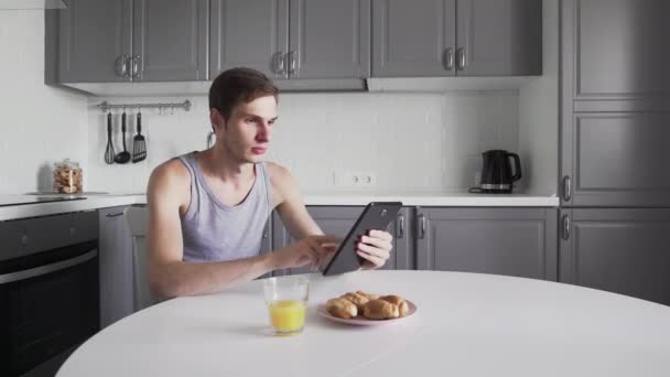 Hombre charlando en la computadora de la tableta y beber jugo por mesa en la cocina — Vídeo de stock