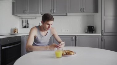 Genç adam portakal suyu içiyor ve elinde akıllı bir cihaz kullanıyor. Sabahları modern mutfakta oturan yakışıklı adamın günlük rutini.