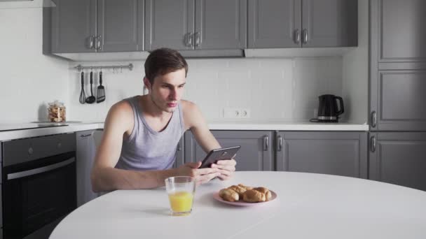 Hombre joven usando tableta digital para leer noticias mientras está sentado en la cocina en casa — Vídeo de stock