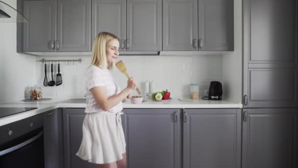 Glad aktiv rolig ung kvinna håller trä spatel mikrofon sjunga karaoke sång dans lyssna musik matlagning i köket — Stockvideo