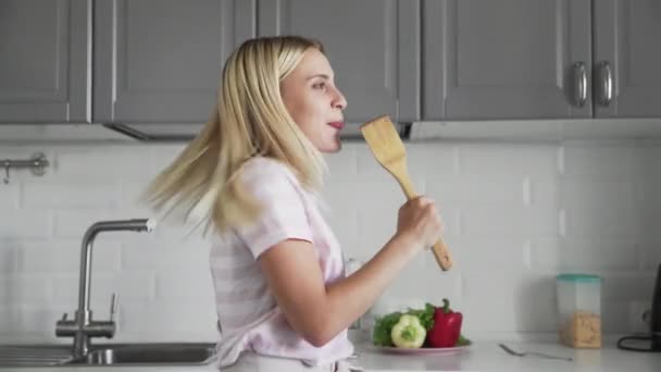 Close up of happy active funny young woman holding wooden spatula microphone singing karaoke song dancing listening music cooking in kitchen — Stock Video