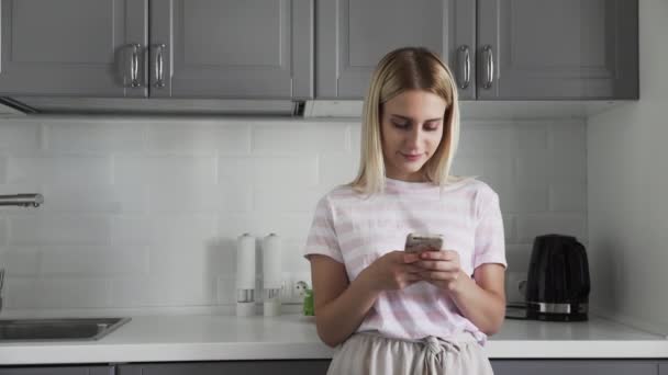 El primer plano de la muchacha joven por la mañana en la cocina que comprueba teléfono. Mujer charlando con amigos usando un teléfono móvil. Chica escribiendo un mensaje en el teléfono inteligente . — Vídeo de stock