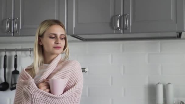 Jolie fille dans une couverture à carreaux boit du thé et regarde par la fenêtre dans la cuisine le matin — Video