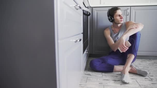 Молодой человек слушает музыку на смартфоне в наушниках, танцует, сидит на полу на кухне и смотрит в окно — стоковое видео