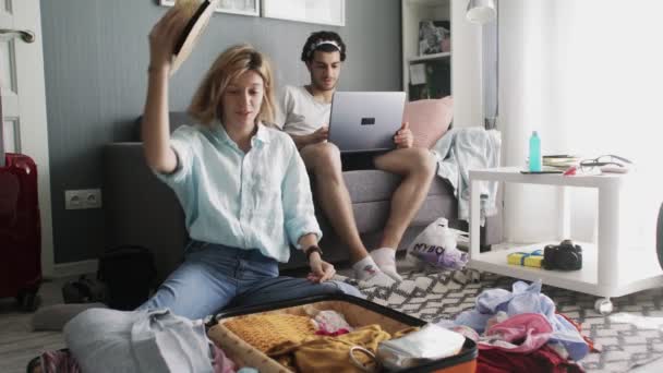 Cara com um laptop no sofá e a menina no chapéu colocar roupas em uma mala e prepare-se para viajar — Vídeo de Stock