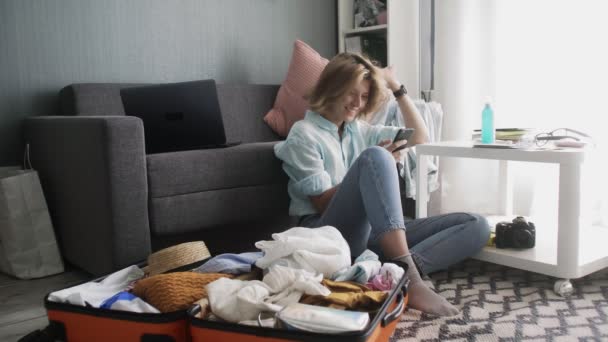 A young girl near a travel suitcase looks at social networks on the phone, laughs and writes with friends — Stock Video