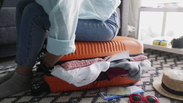 Милая девушка, сидящая на переполненном чемодане, пытается закрыть его — стоковое видео