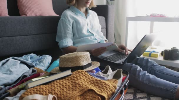 Ung flicka med bärbar dator nära resväska, gör sig redo för resa och kontrollerar semesterbiljetter — Stockvideo