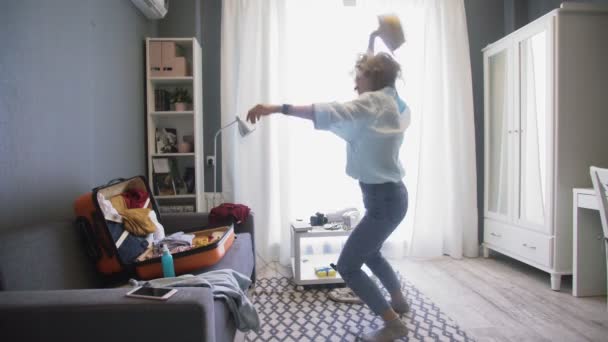 Mujer joven feliz escuchando su música favorita y bailando mientras empaca su bolso de viaje en casa o en la habitación de hotel . — Vídeos de Stock