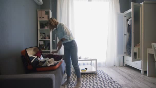 Молодая женщина берет одежду из шкафа и кладет ее в чемодан — стоковое видео