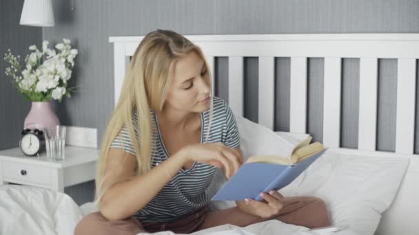 Mujer joven leyendo libro y hablando por teléfono en la cama en casa — Vídeos de Stock