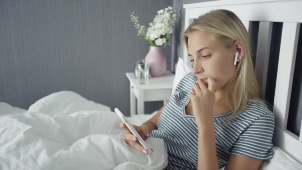 Glückliche schöne junge Frau mit drahtlosen Kopfhörern, die auf dem Bett liegen und das Smartphone benutzen — Stockvideo