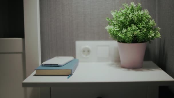 Female hand turn off an alarm clock on mobile phone on bedside table with indoor plant and book — Stock Video