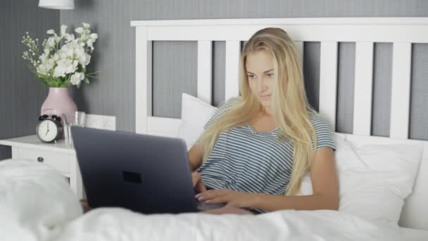 Жінка сидить на ліжку і друкує на блокноті. Молода біла жінка з комп'ютером вдома. Чарівна дівчина фрілансер, що працює на ноутбуці вдома — стокове відео