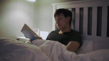 Genç Asyalı adam yatakta uzanıp kitap okuyor.