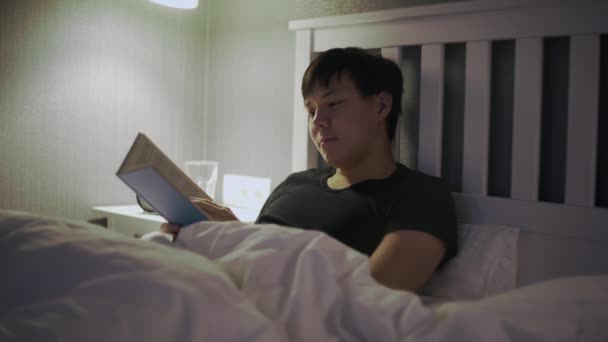 Joven asiático hombre acostado en la cama y leyendo un libro — Vídeos de Stock