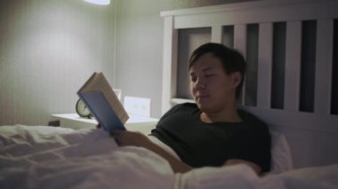 Genç Asyalı adam yatakta uzanıp kitap okuyor.