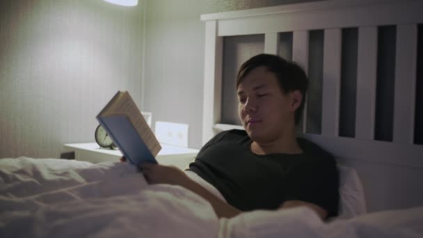 Νεαρός Ασιάτης ξαπλωμένος στο κρεβάτι και διαβάζοντας ένα βιβλίο — Αρχείο Βίντεο